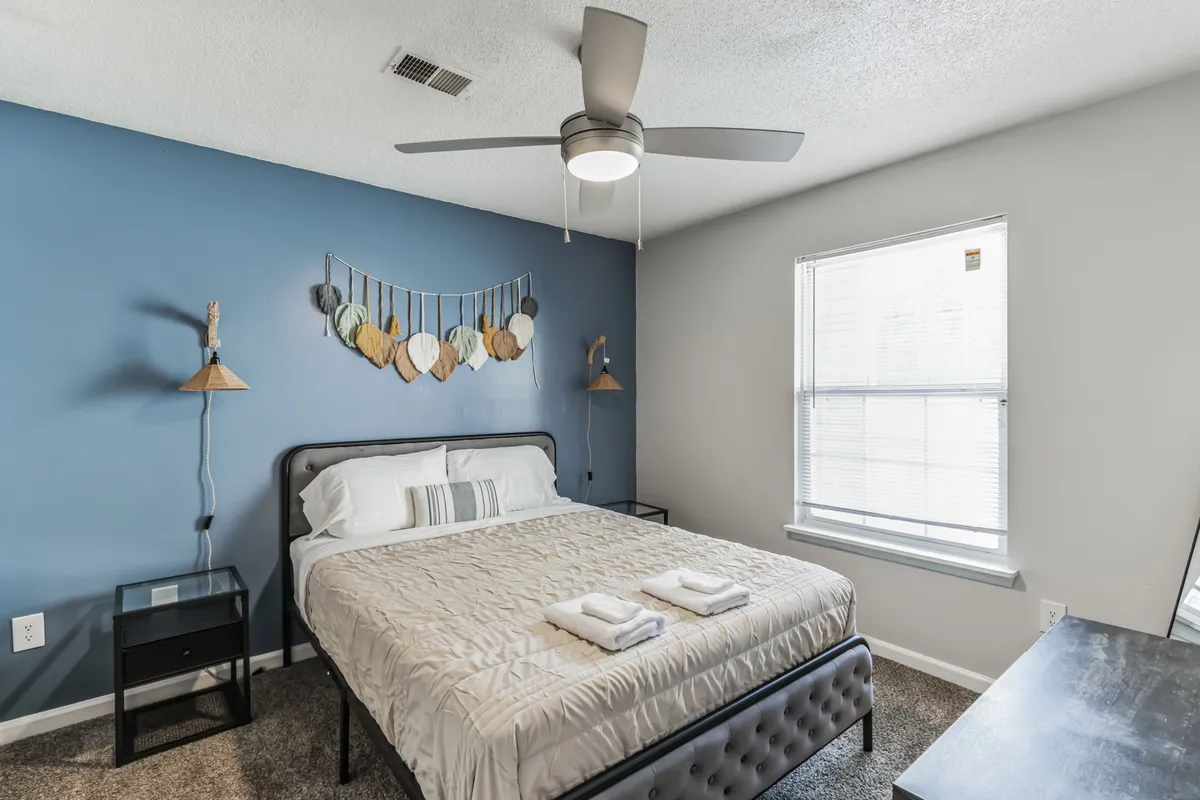 The image showcases a modern, sunlit bedroom featuring a plush queen bed with crisp linens, stylish decorative wall art above the headboard, matching nightstands with soft lighting, and a large window that fills the space with natural light. The overall atmosphere is cozy and inviting, ideal for relaxation.