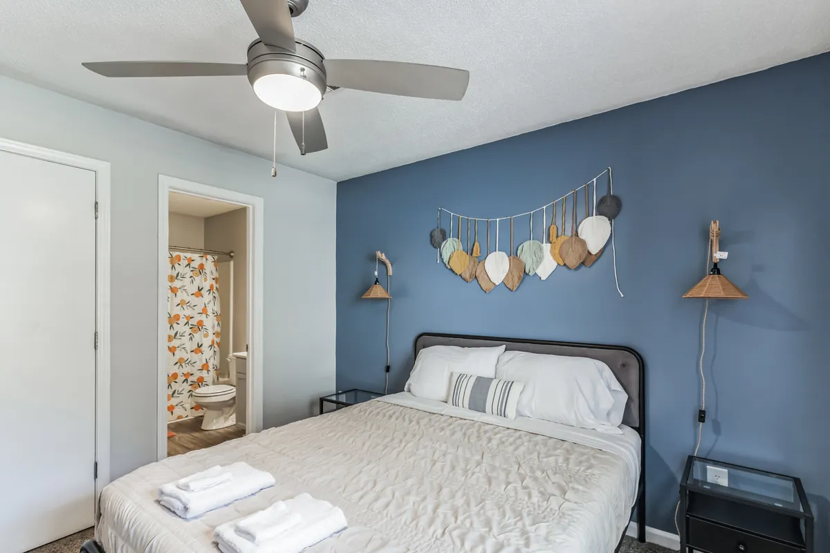 A serene bedroom interior featuring a plush queen bed with crisp white linens, a decorative leaf wall hanging above the headboard, and a ceiling fan. Soft, natural light filters in, creating a peaceful and cozy atmosphere ideal for restful sleep.