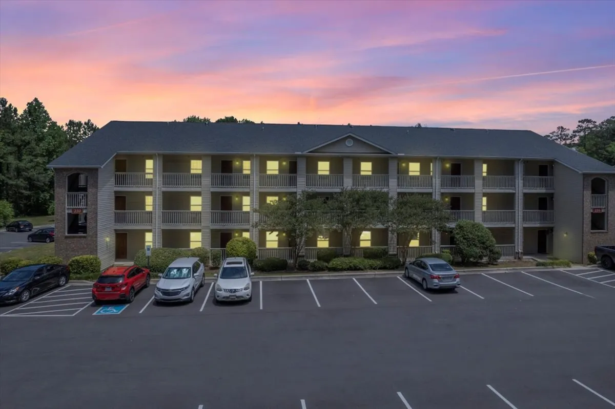 This image showcases the apartment building's welcoming facade, with softly illuminated windows and a lively parking area where residents are seen relaxing on balconies or strolling through the grounds. The scene exudes a sense of community and comfort, ideal for residents seeking a safe and convenient home base close to Fort Bragg.