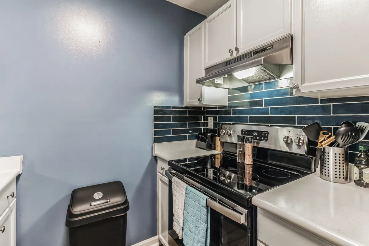This image features a bright, modern kitchen with stainless steel stove, blue backsplash tiles, and crisp white cabinetry. Kitchen accessories are visible on the counters, suggesting a space ready for meal preparation or casual cooking. The scene exudes a sense of functionality paired with a welcoming, homey atmosphere.