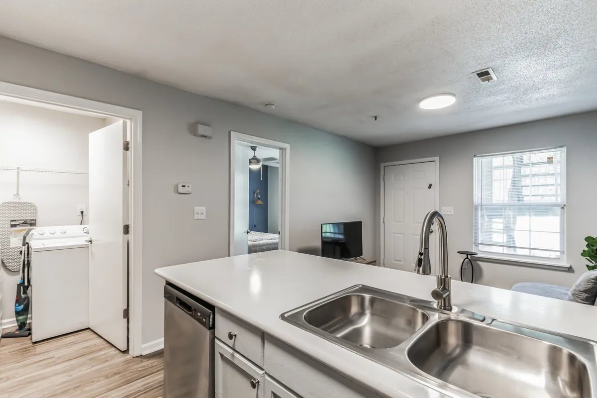 A spacious, comfortable kitchen interior that subtly includes the washer and dryer alongside the kitchen island and sink. Natural light filters in through a window, and the scene conveys a lived-in, multifunctional space where cooking, socializing, and laundry can happen seamlessly.