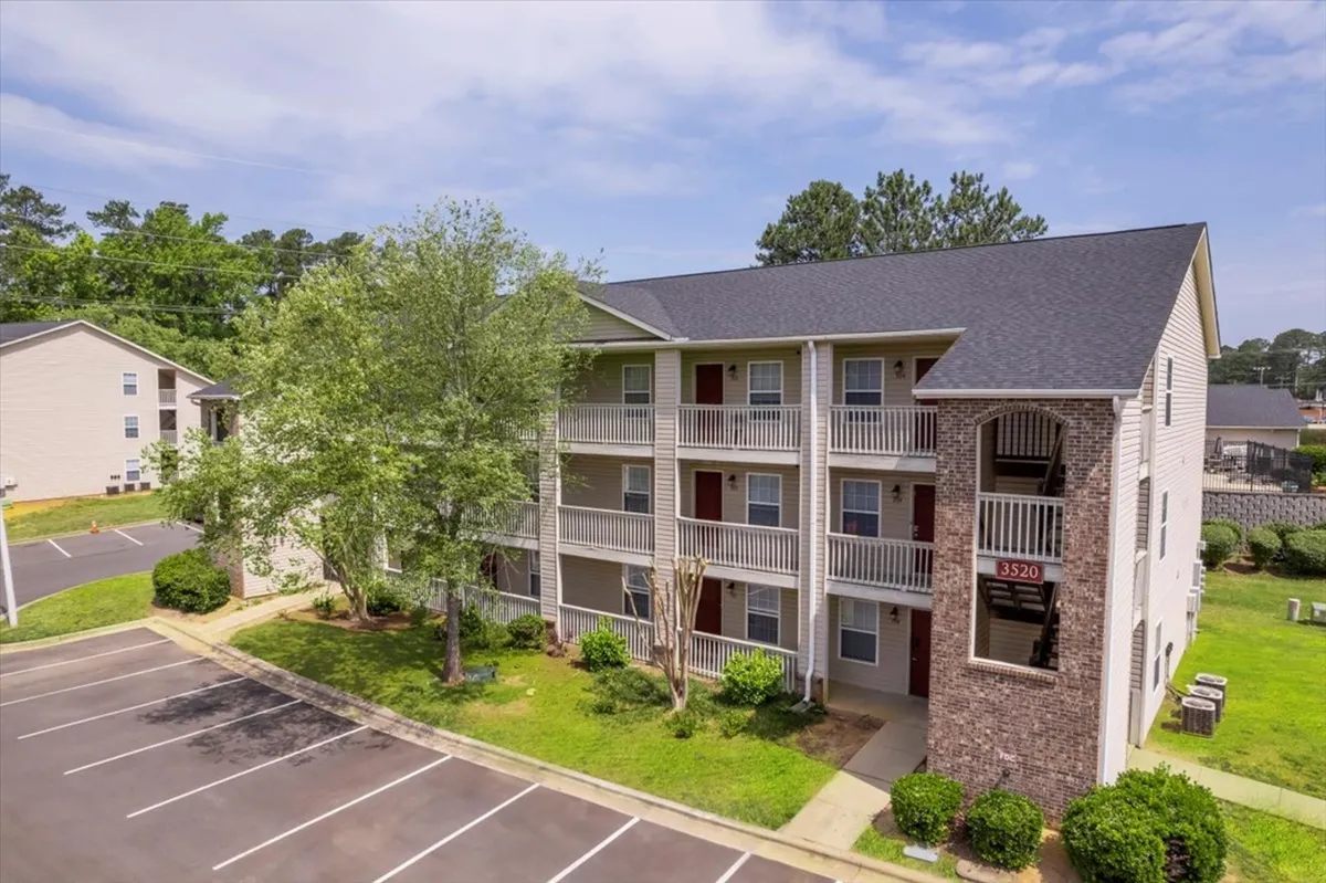 A welcoming exterior view of a three-story apartment building surrounded by lush green landscaping and a well-maintained parking lot. The scene suggests a comfortable and accessible community environment ideal for business travelers seeking convenience and a sense of home.