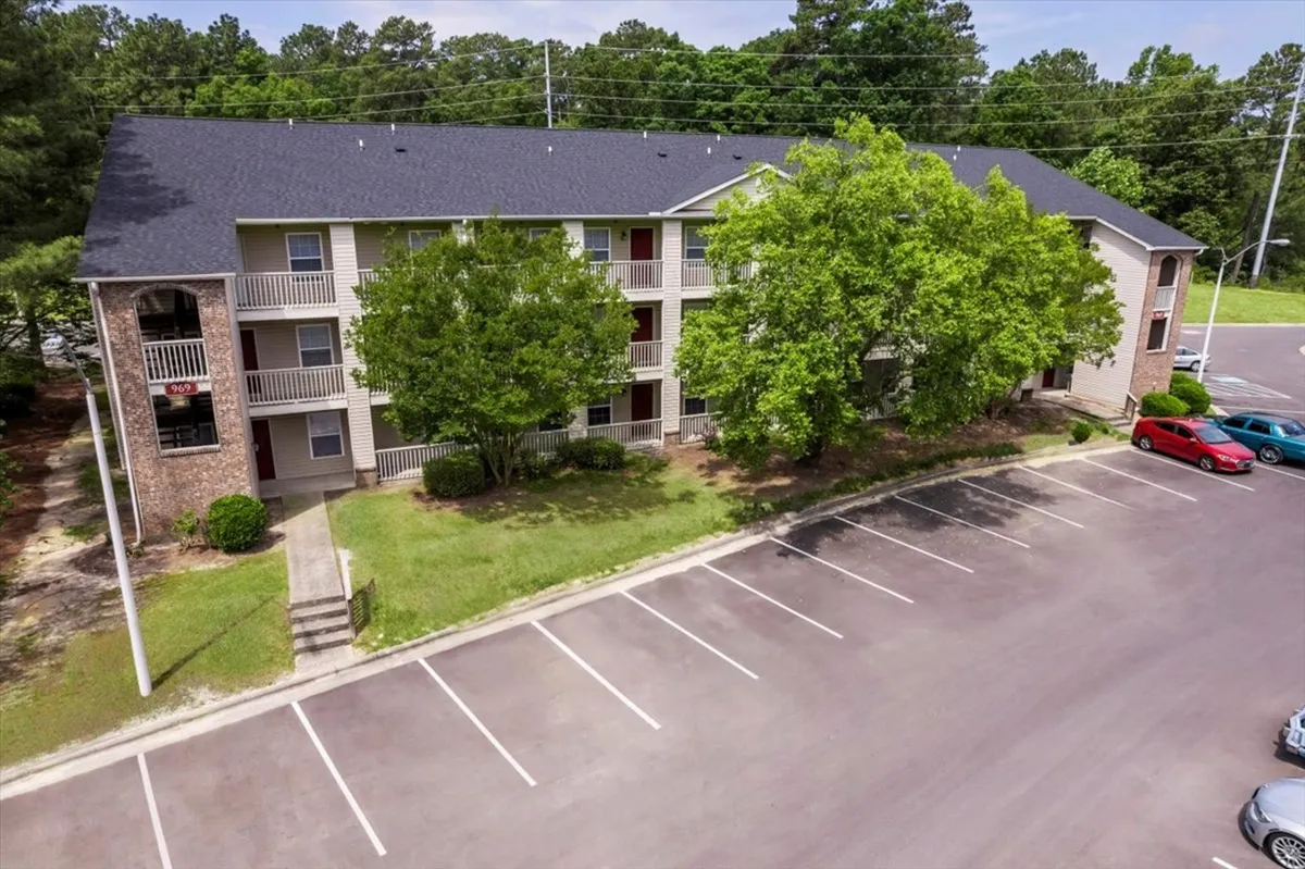 A sunlit exterior perspective capturing a large green tree, the building’s clean facade, and the adjacent parking area. The inviting greenery and outdoor space hint at the property's proximity to amenities and relaxing spots for breaks during a busy stay.
