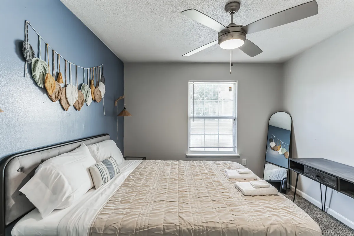 A tranquil bedroom scene with a bed dressed in neutral bedding, a decorative wall hanging, a mirror, and a window letting in gentle natural light. A black desk stands nearby, offering additional utility for reading or getting ready.