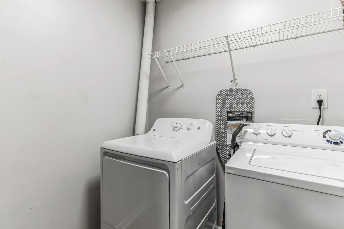 A dedicated laundry area featuring a modern washing machine and dryer neatly positioned side by side, with a convenient shelf above for detergent and laundry supplies. An ironing board is tucked to the side, creating a practical and efficient space for handling laundry tasks.